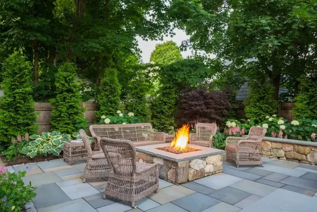 Outdoor patio with a stone fire pit and wicker chairs arranged around it, set on a tiled surface amid lush greenery and flowering shrubs