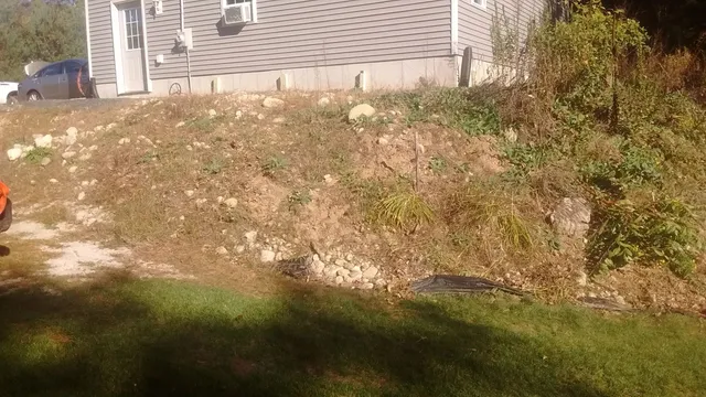 A residential property in Dudley before landscaping, showing a steep, rocky slope with dry grass and scattered stones leading up to a house with light gray siding. The terrain is unlandscaped and uneven.