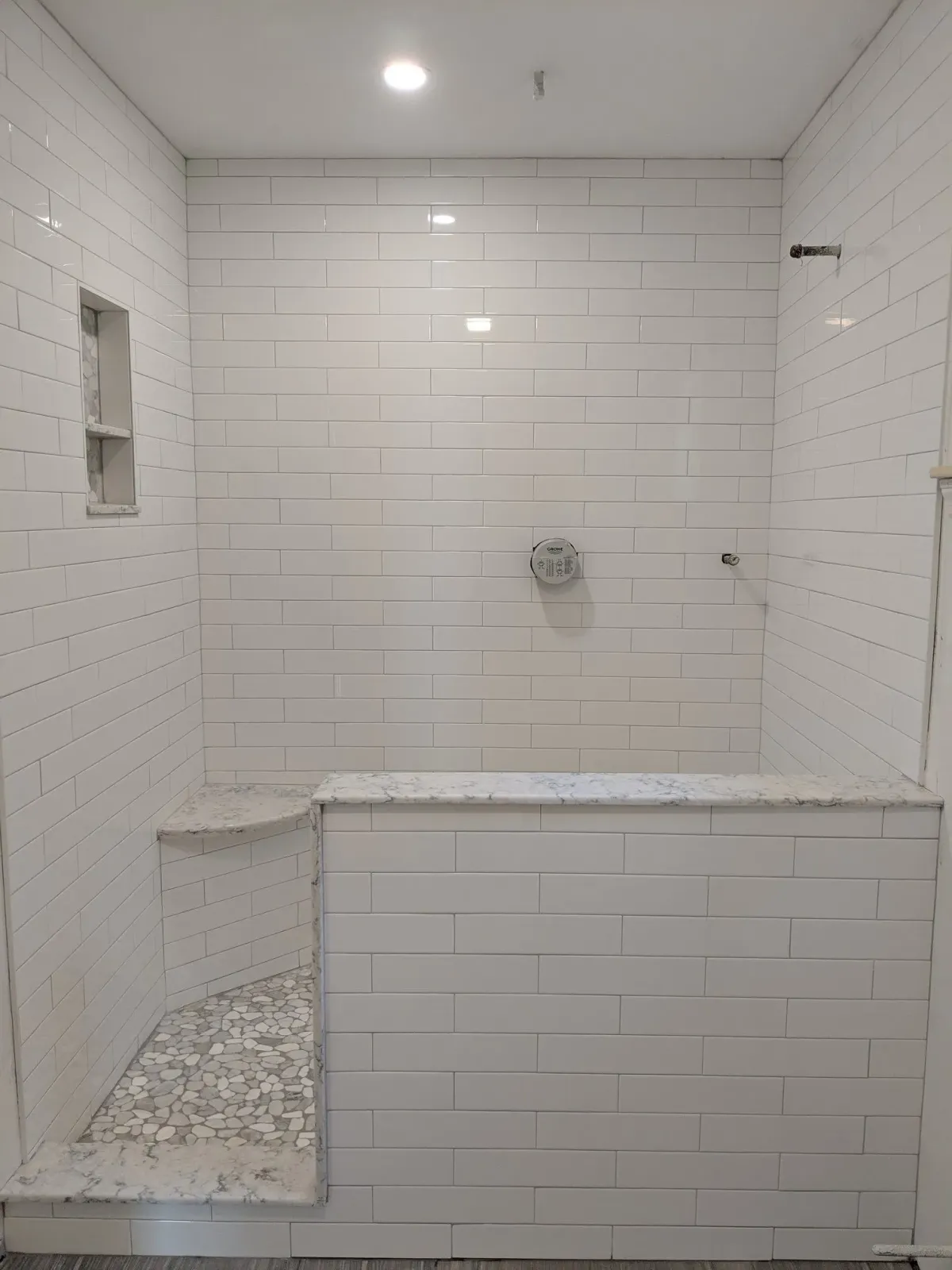 White tiled shower stall with built-in bench and pebble floor area, with a niche and shower fixtures.