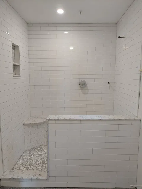 White tiled shower stall with built-in bench and pebble floor area, with a niche and shower fixtures.