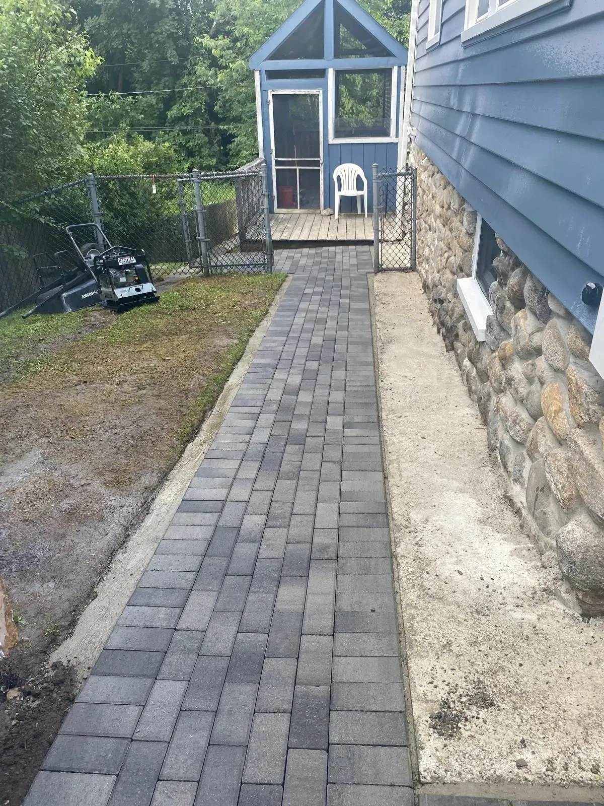 Narrow paver walkway beside a blue house leads to a screened porch and patio chair in the backyard.