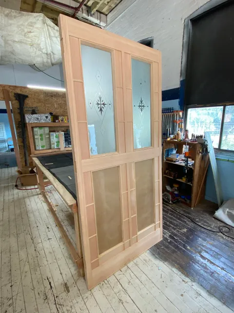 A partially assembled wooden cabinet with glass panels and decorative designs in a workshop. There are tools and materials scattered around on shelves and workbenches, with a window letting in natural light.