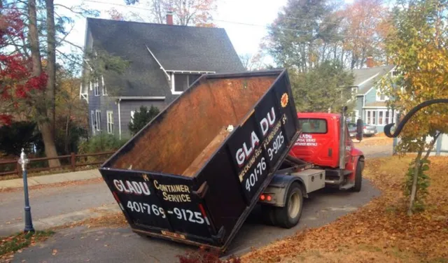 A red truck with an open black container bed tilted upward parked on a residential street during autumn, with fallen leaves and houses in the background.