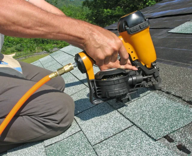 Person kneeling on roof using pneumatic nail gun to secure shingles.