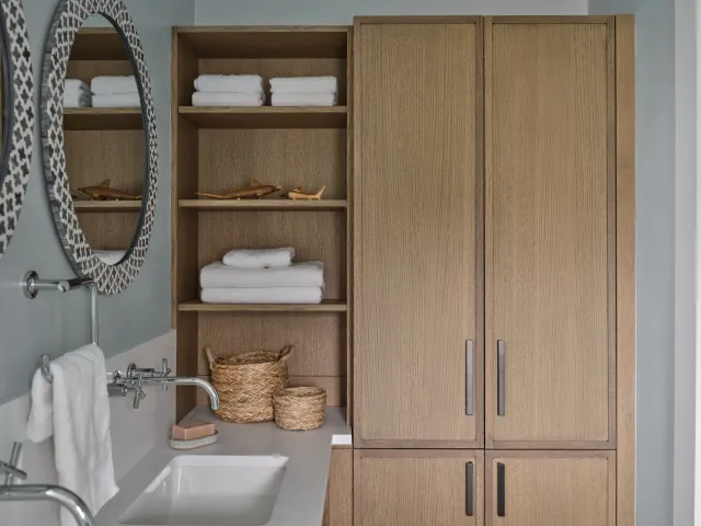 Bathroom with wooden cabinets, shelves holding folded towels and wooden toy sharks, oval mirrors with decorative frames, a white sink, soap, and a towel hanging on a silver towel rack.