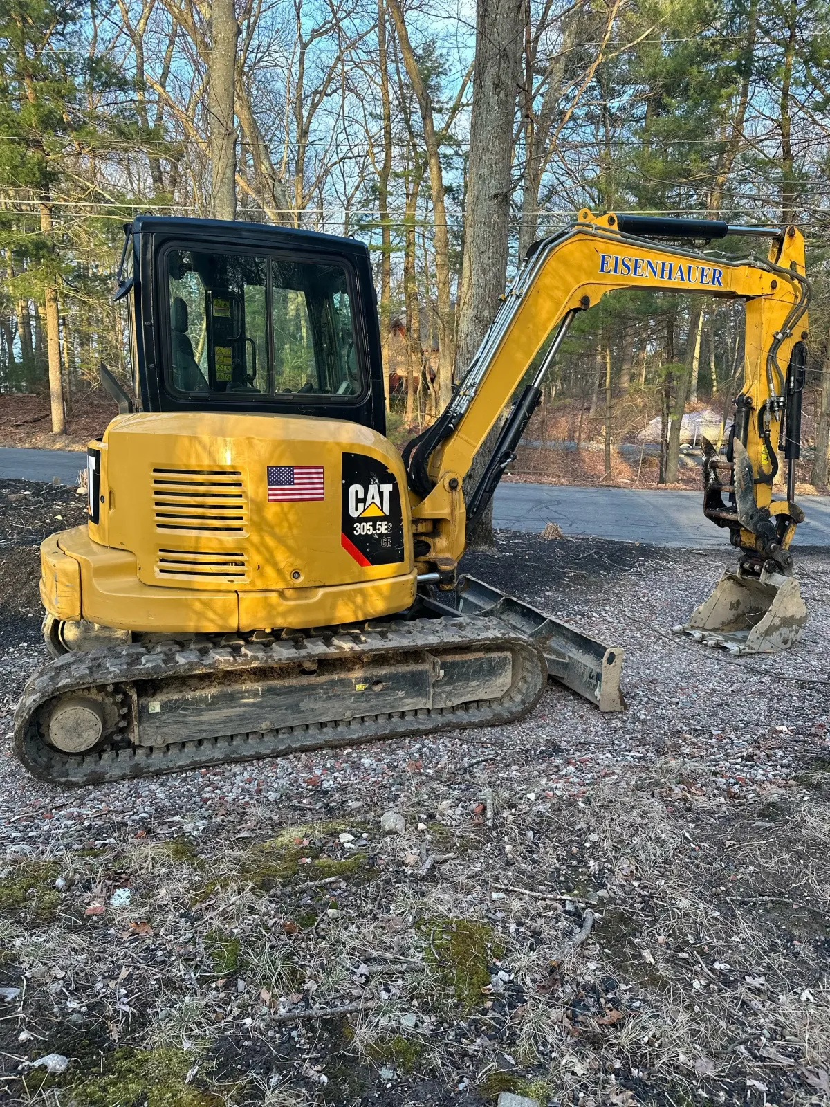 A yellow CAT mini excavator with the brand name EISENHAUER on the arm, sitting on a patch of dirt and gravel in a wooded area with trees and a road in the background.