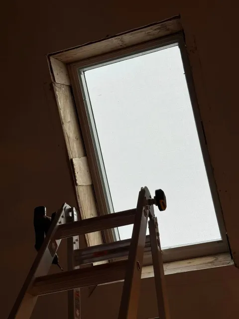 A ladder angled towards a rectangular skylight in a brown ceiling.