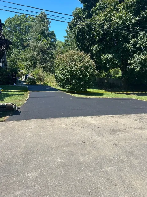 Paved driveway leading into a tree-lined area. The driveway is freshly paved with a dark color.
