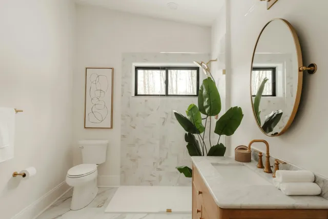 Cypress Works Massachusetts, a modern bathroom featuring a round mirror, a marble countertop with a bronze faucet, a potted plant, a framed abstract line drawing, a toilet, and a window with a view of trees outside.