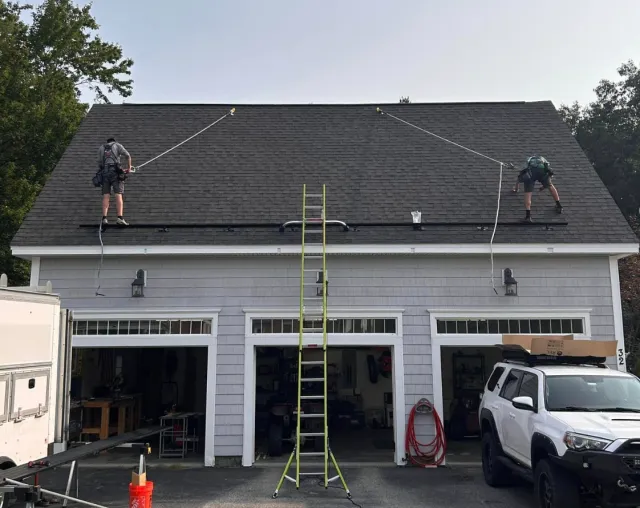 Two workers are on the roof of a house, cleaning or inspecting the shingles using ropes. A ladder is set up against the house, and a garage with open doors is visible below, with various tools and equipment inside. A white SUV is parked in front of t