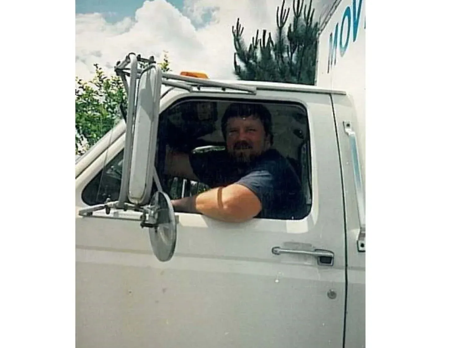 A man with a beard and dark hair driving a white truck, looking out the window with his arm resting on the windowsill, under a partly cloudy sky.