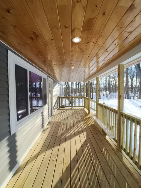 New wooden porch with railing attached to house, with snow-covered trees in the background and a clear sky.