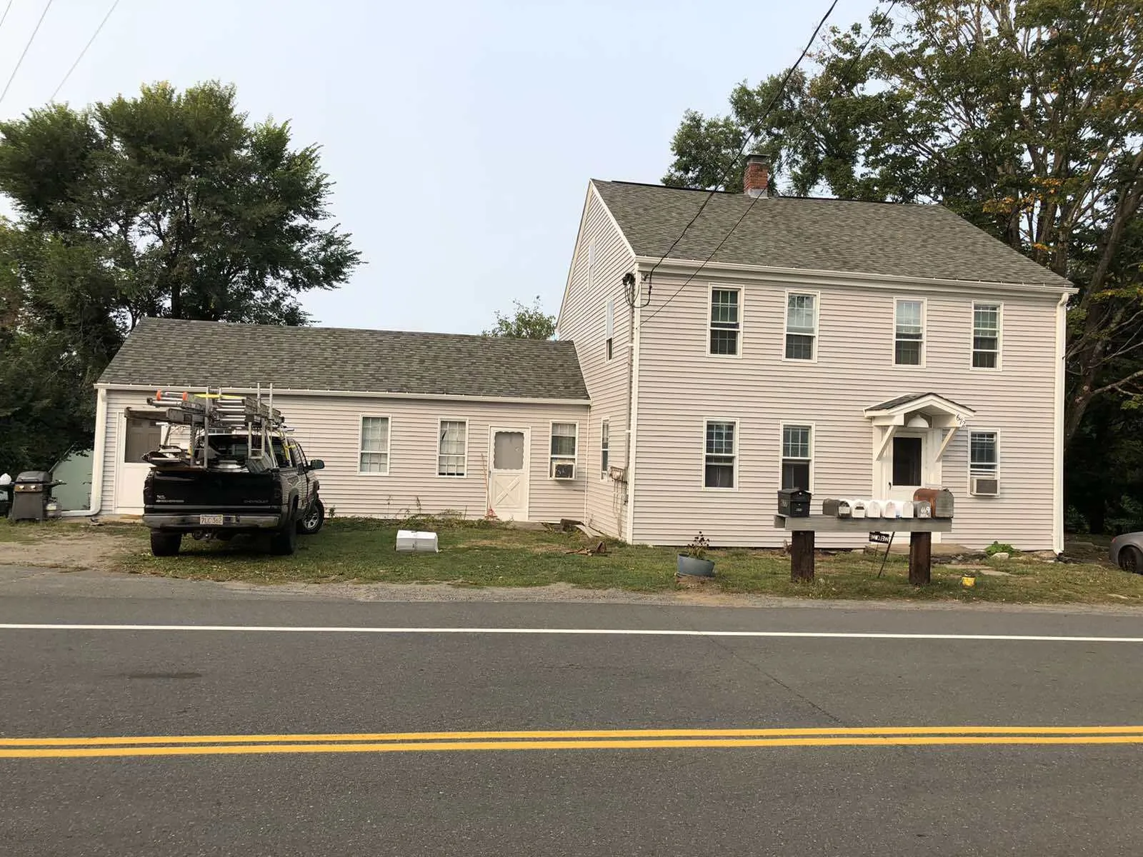 A two-story white house with an attached single-story section and a pickup truck parked in front, beside a two-lane road.