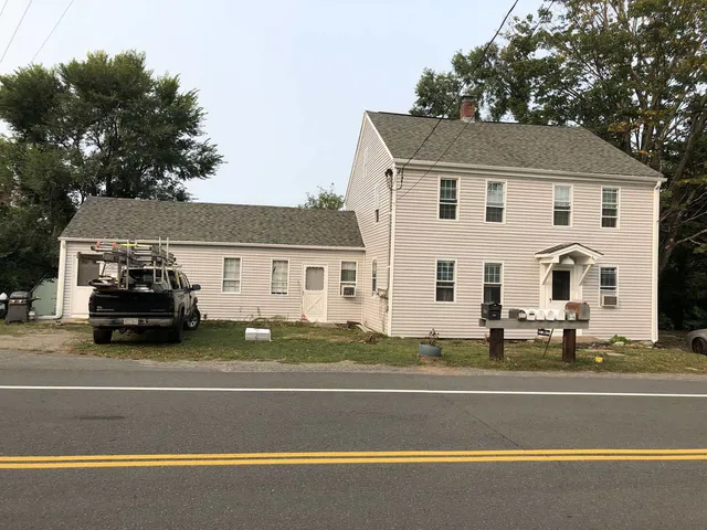 A two-story white house with an attached single-story section and a pickup truck parked in front, beside a two-lane road.