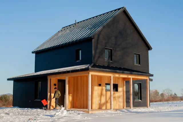 A two-story house with black siding and a metal roof, with part of the front porch unfinished with exposed wood beams, and a man shoveling snow outside on a snowy landscape under a clear blue sky.
