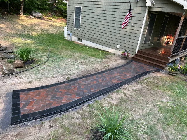 Brick walkway with black border leading to a house porch, next to an American flag and lit steps.