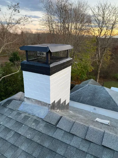 A white painted brick chimney with a black metal cap, situated on a shingled roof against a backdrop of trees at dusk.