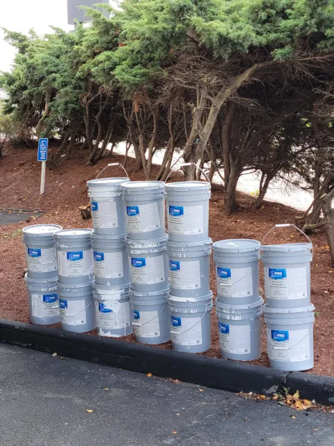 Multiple buckets of paint stacked in front of trees in a parking lot, with a disability parking sign nearby.
