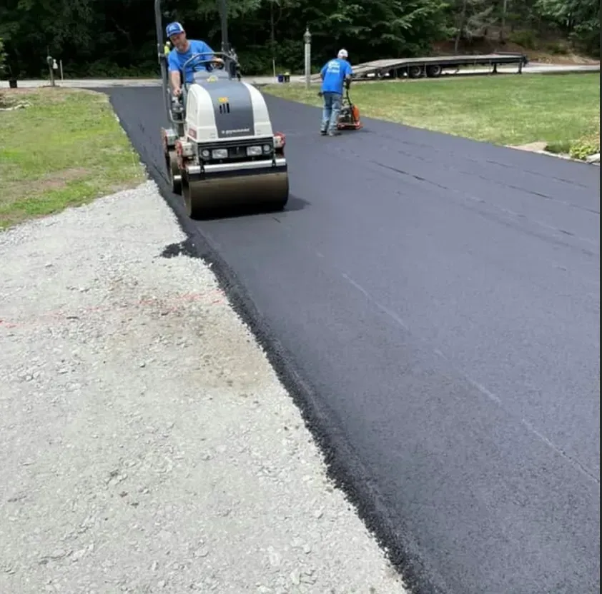 Construction workers use a steamroller and hand-operated compactor to smooth fresh asphalt on a driveway.