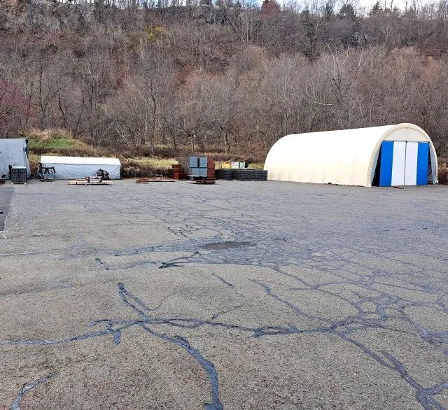 A gravel lot with a large, arched white storage tent and scattered equipment, bordered by trees on a hillside.