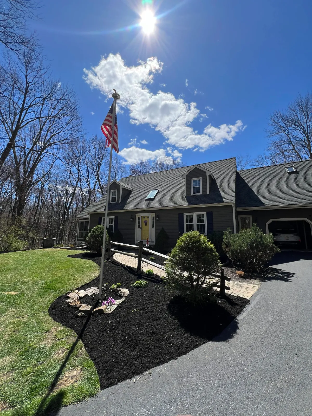 A gray house with a landscaped front yard, American flag on a pole, bright sunny sky with some clouds, leafless trees in the background, and a paved driveway.