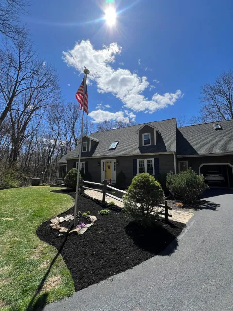 A gray house with a landscaped front yard, American flag on a pole, bright sunny sky with some clouds, leafless trees in the background, and a paved driveway.