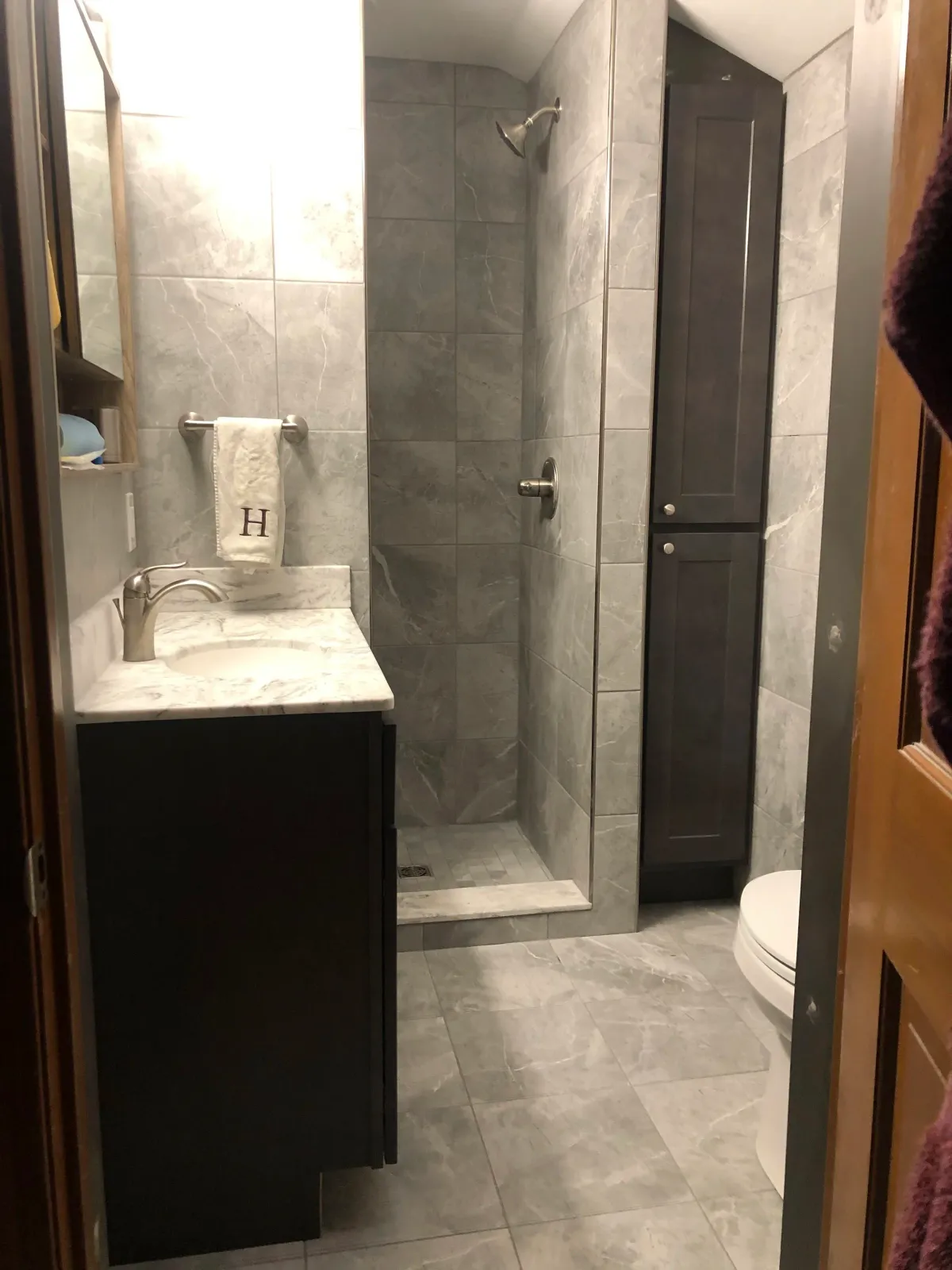 A small modern bathroom with gray marble tiles on the floor and walls. It features a walk-in shower with a glass door, a white towel with an embroidered letter 'H' hanging on a towel rack, a dark vanity with a marble countertop, a sink, and a faucet. There is a tall dark storage cabinet next to the shower and a white toilet visible in the corner.