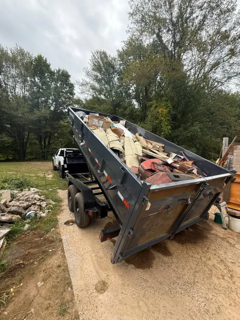 Dump truck with bed raised, full of debris, backed up on a dirt driveway next to a grassy area with trees under an overcast sky.