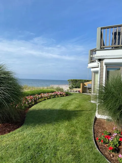 Well-maintained green lawn with flower beds beside a beach house overlooking the ocean under a partly cloudy sky.