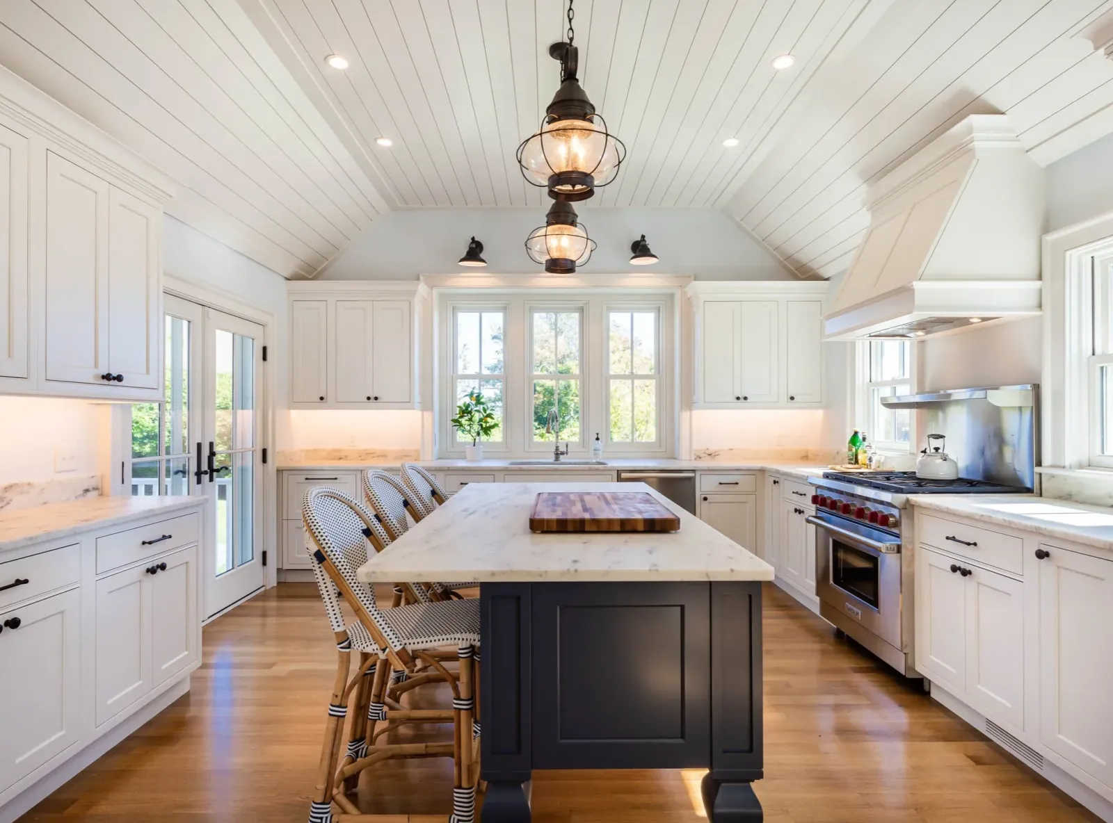 Kitchen with white cabinets, a marble island, a black kitchen island table, hanging pendant lights, windows with a view of greenery outside, and a wooden floor.