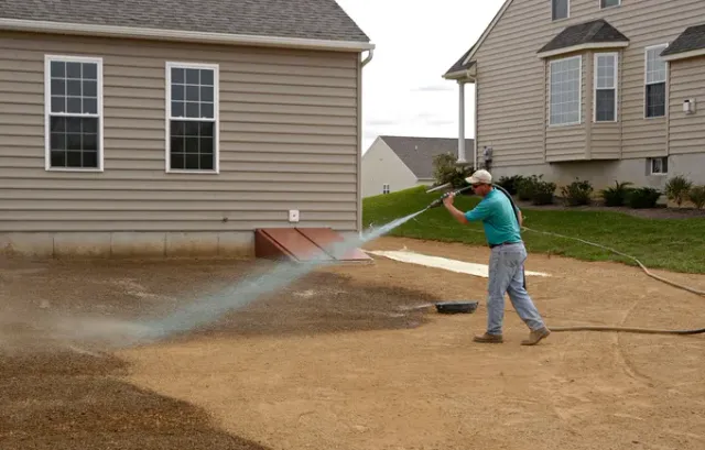 Person spraying water from a hose across a dirt yard beside suburban houses.