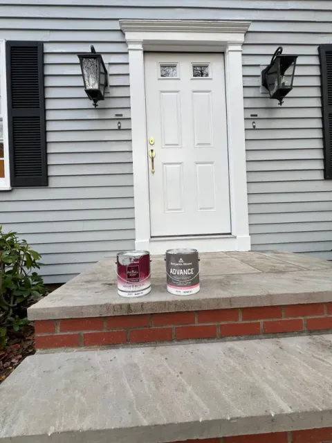 Two paint cans, one dark red and one grey, sit on the brick and stone front steps of a house with a white door.