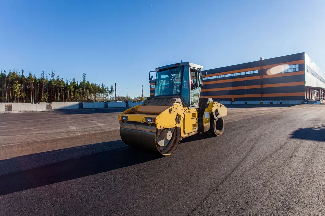 A yellow road roller compacts freshly laid asphalt on a construction site next to a building under a clear blue sky.