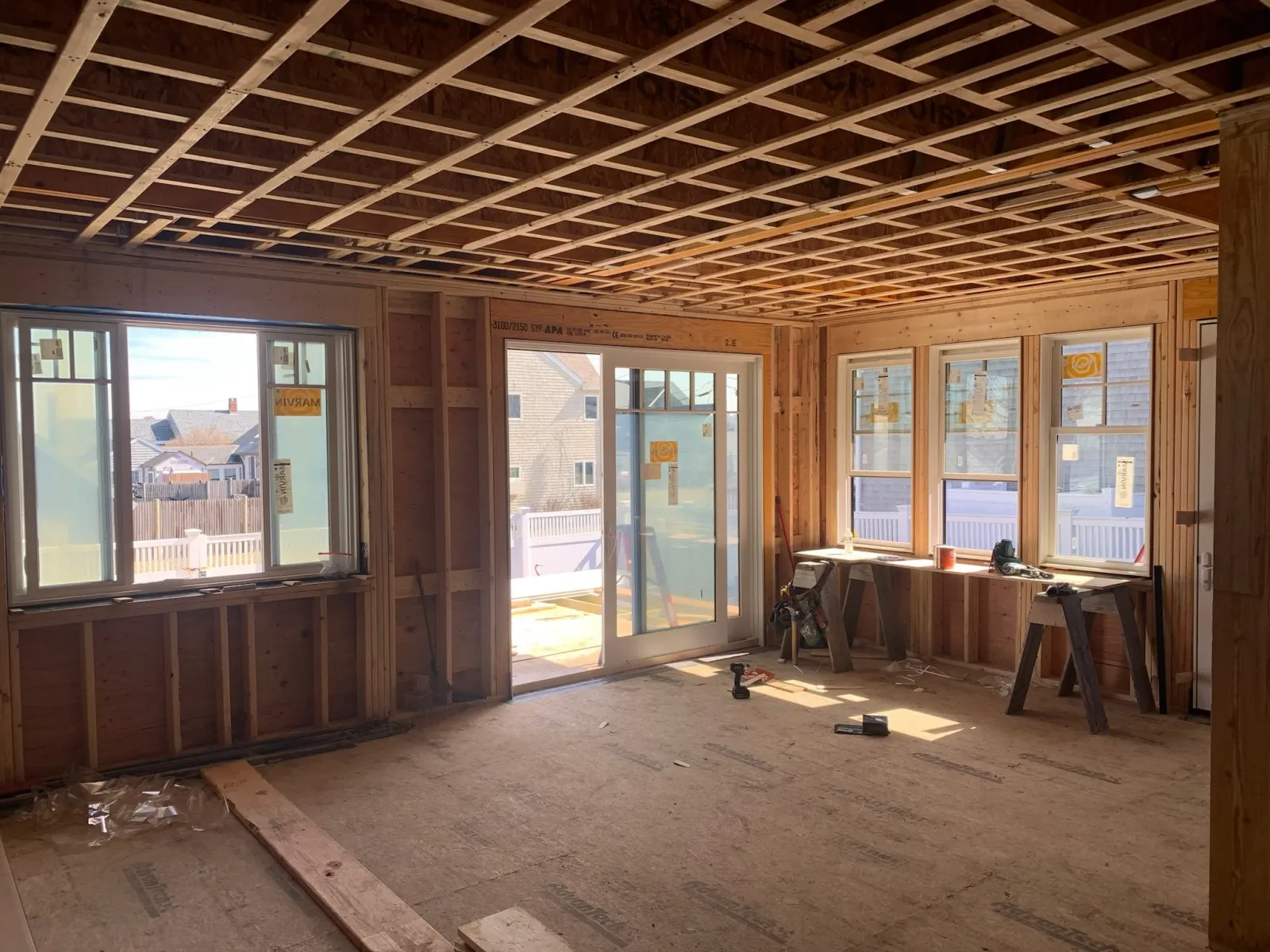 Interior view of a house under construction with unfinished wooden framing, multiple windows, and a sliding glass door leading outside. Construction tools and materials are scattered on the floor. Scituate, Norwell, Hanover, Hanson, Marshfield.