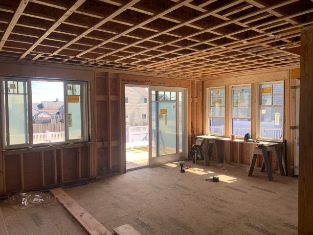 Interior view of a house under construction with unfinished wooden framing, multiple windows, and a sliding glass door leading outside. Construction tools and materials are scattered on the floor. Scituate, Norwell, Hanover, Hanson, Marshfield.