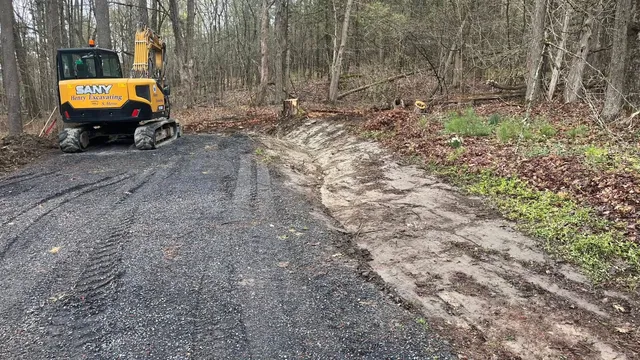Yellow excavator on a gravel road beside a wooded area, working near a muddy shoulder.