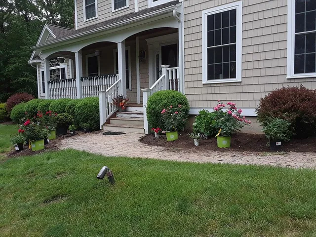 Front porch of a gray house with potted flowers, white railings, and a green lawn in front