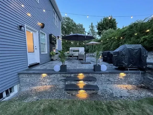 Backyard patio with string lights, stone steps, and grill, at dusk.