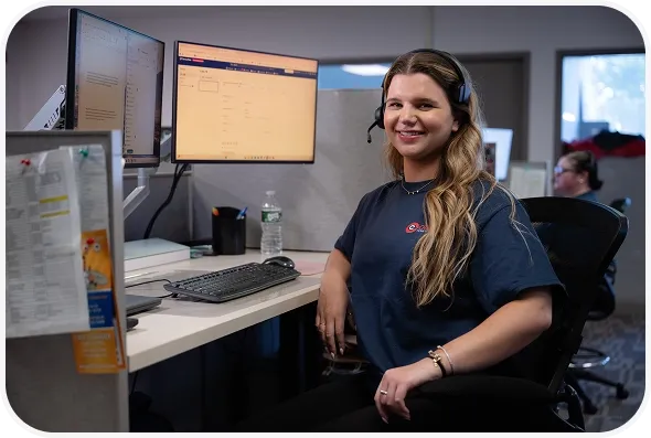 A friendly Cranney Home Services team member with a headset smiles at her dual-monitor desk; a coworker works in the background.