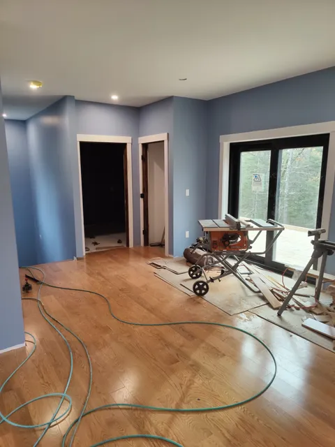 Room under renovation with blue walls, hardwood floor, and construction tools. A table saw is set up near a large window, and electrical cords are scattered on the floor.