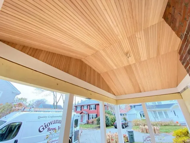 View of a porch ceiling made of wooden planks with a section of brick wall in the corner and a neighborhood street with houses and a van in the background.