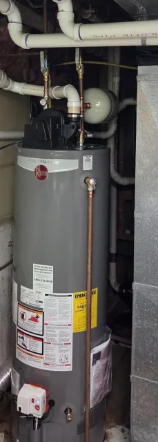 A Rheen Performance water heater system with copper and PVC pipes connected to it, situated in a utility room. Pipes are labeled with blue and red, indicating cold and hot water lines. The heater has warning and energy guide labels.