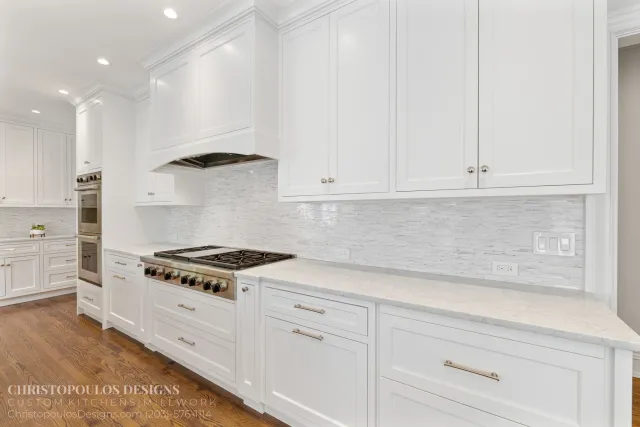 White kitchen with cabinets, a built-in oven, stainless steel stove, and a light-colored backsplash. Hardwood flooring and recessed ceiling lights.