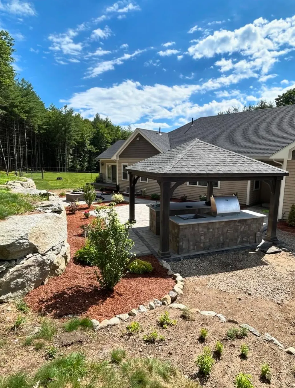 Outdoor patio with a wooden pergola and stone grill station, surrounded by landscaped garden, rocky border, and a backdrop of trees and blue sky.