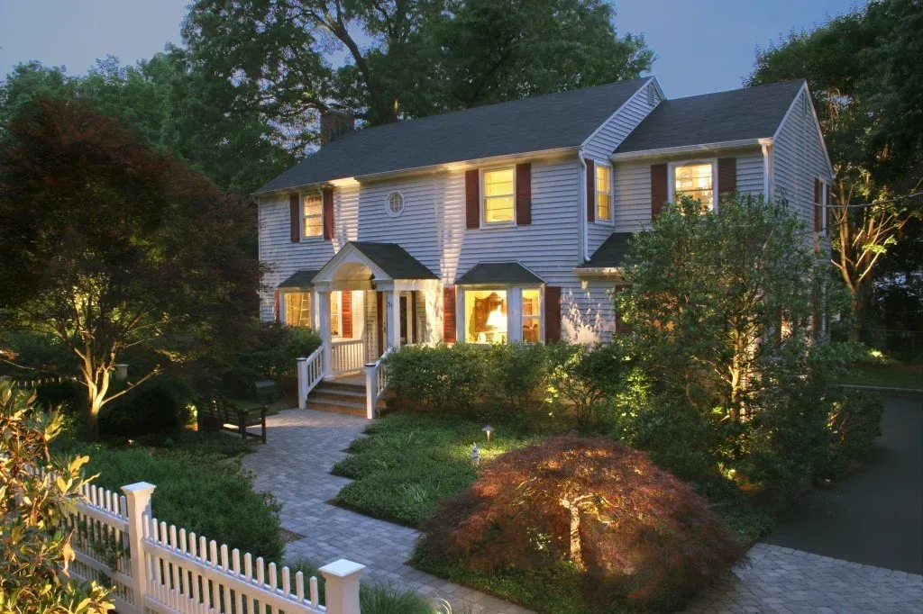 Two-story white house with porch, surrounded by greenery and illuminated at dusk.
