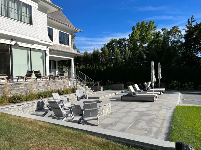 Backyard patio with lounge chairs, umbrellas, and a fire pit next to a swimming pool, with a large white house and trees in the background.