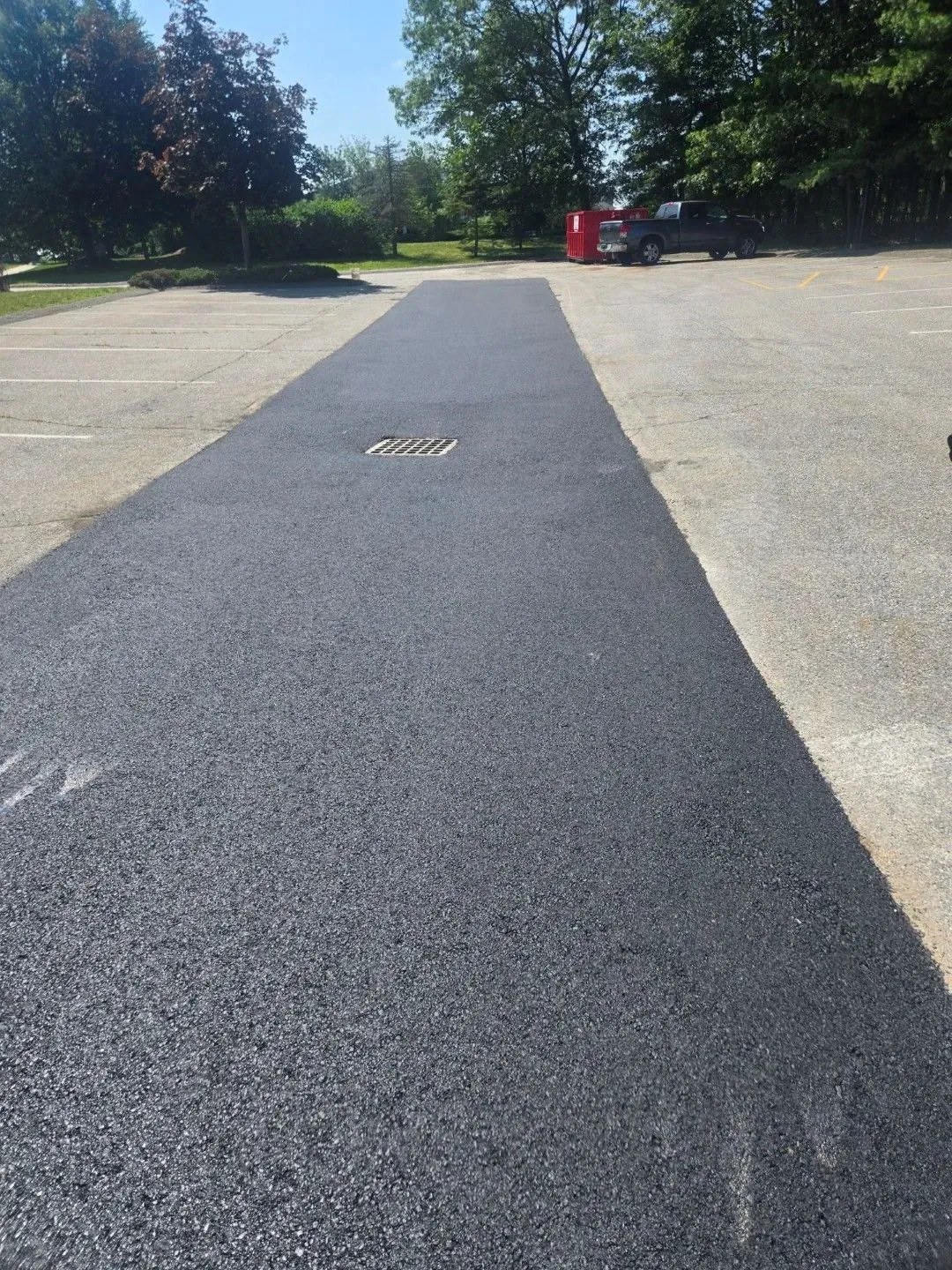 Dark paved path across a gravel lot with trees in the background and a red bin near the end.