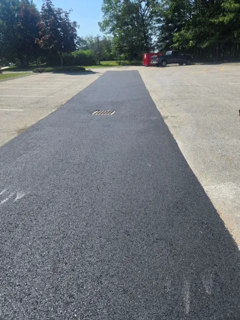 Dark paved path across a gravel lot with trees in the background and a red bin near the end.