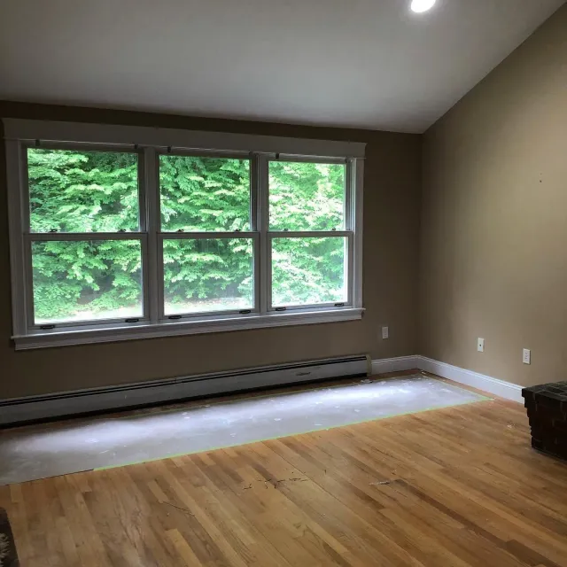 Empty room with hardwood floor, large windows, beige walls, and greenery outside.