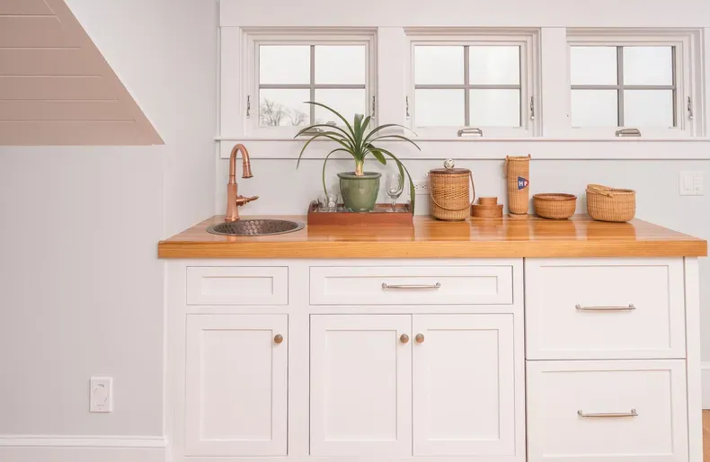 Bright kitchen counter with white cabinets, wooden countertop, sink, plants, and woven storage baskets.